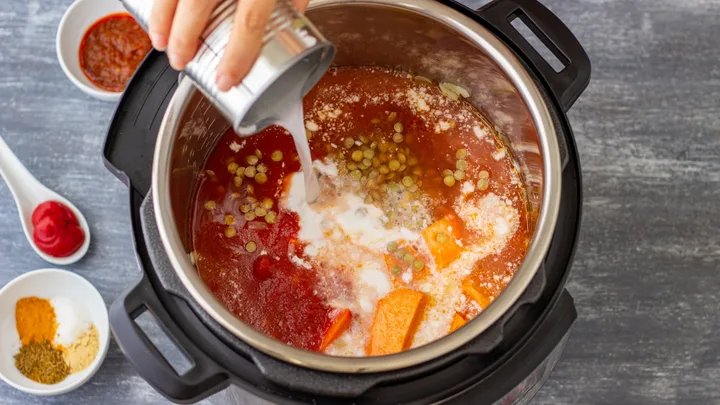 Electric pressure cooker viewed from above with ingredients inside, a can of coconut milk is being poured in