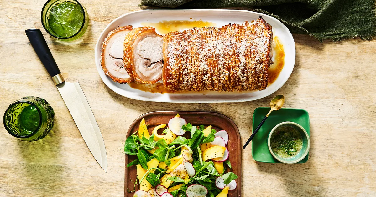 A golden loin of roast pork with thick crackling on top. A sliced mango and snow pea salad is arranged on a shallow platter to the side with a small bowl of nam jim dressing.
