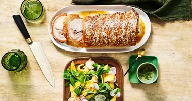 A golden loin of roast pork with thick crackling on top. A sliced mango and snow pea salad is arranged on a shallow platter to the side with a small bowl of nam jim dressing.
