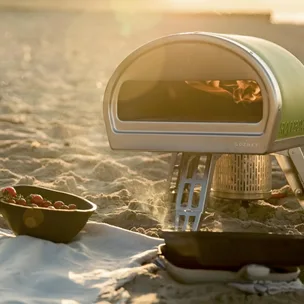 A green Gozney Roccbox portable pizza oven on the beach, cooking a pizza beside a picnic.