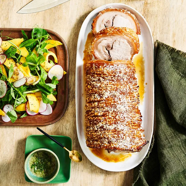 A golden loin of roast pork with thick crackling on top. A mango salad is on the platter to the side with nam jim dressing in a small bowl.