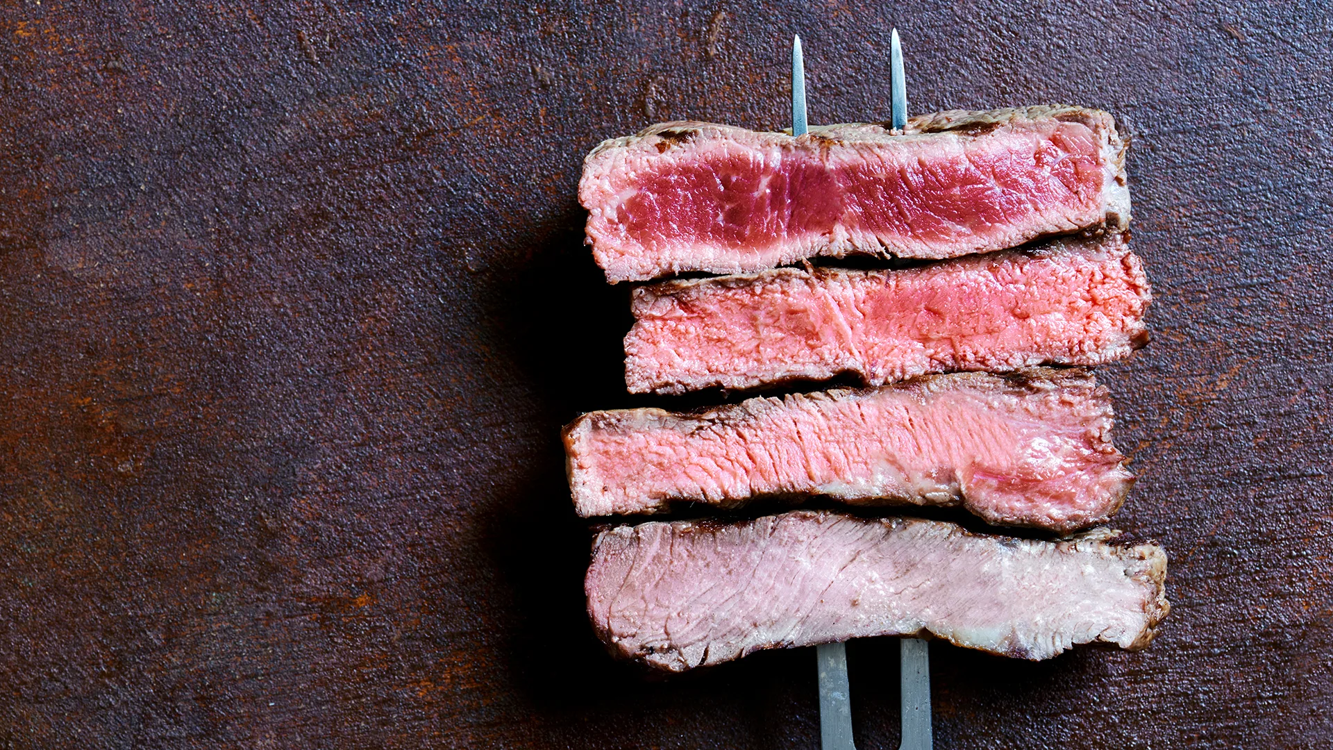 Four pieces of red meat, such as a cut of a steak, cooked and skewered on a fork.