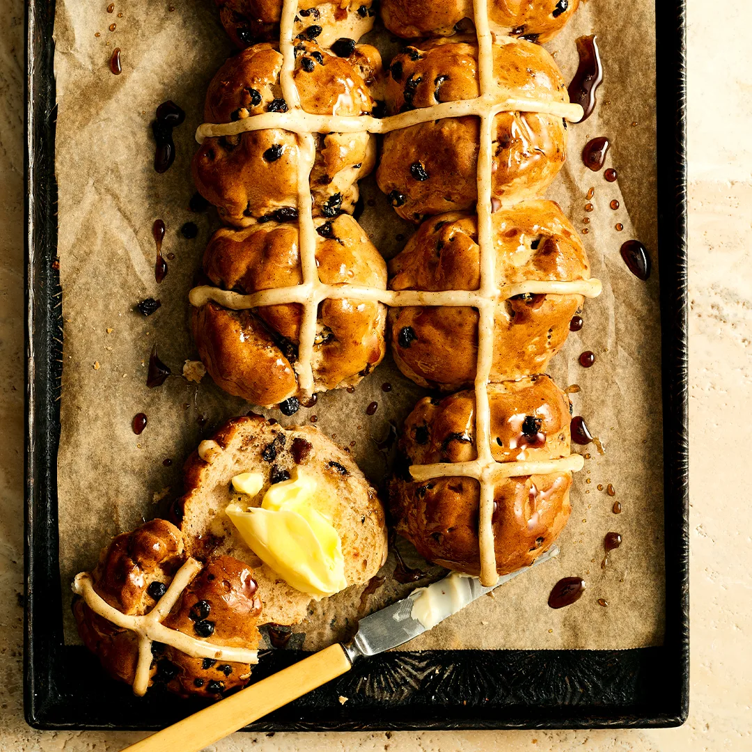 Six gluten free hot cross buns. One has been cut in half and spread with butter, ready to eat.