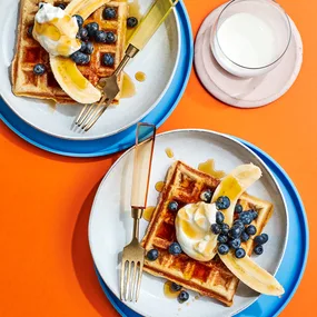 Buttermilk waffle recipe, two waffles with bananas, blueberries and yoghurt, served on white plates with an orange background