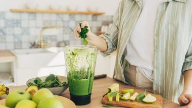 Person adding spinach to a blender with green smoothie ingredients on a kitchen counter.