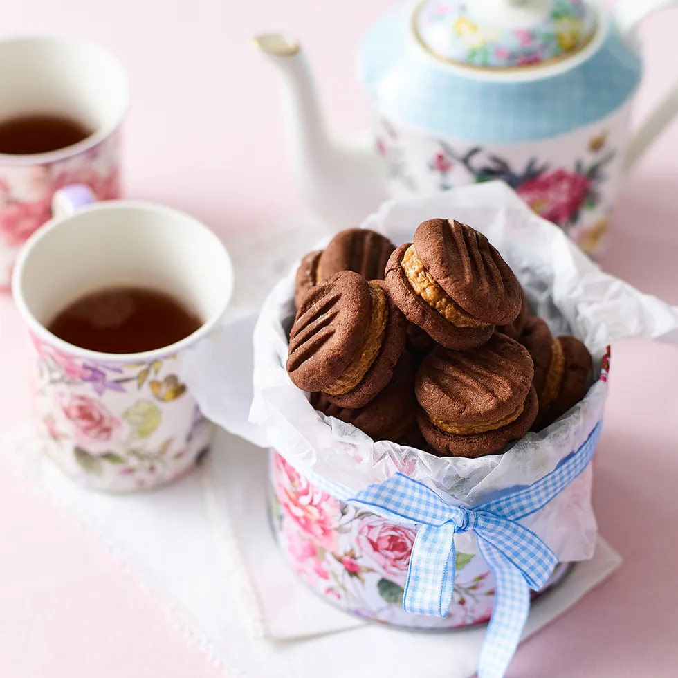 A tin of chocolate Biscoff melting moments next to a teapot and two cups of tea