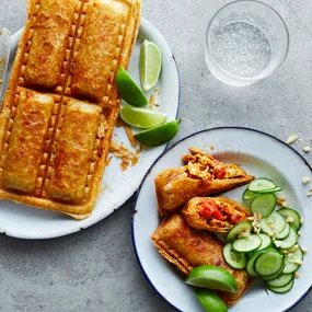 Satay chicken sausage rolls on a plate with cucumber slices, crushed peanuts and lime wedges