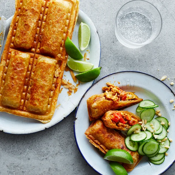 Satay chicken sausage rolls on a plate with cucumber slices, crushed peanuts and lime wedges