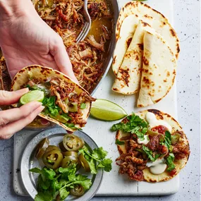 Pulled pork being served from a dish into a tortilla with a squeeze of lime, jalapenos and coriander.