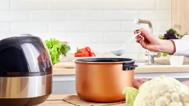 Female hand holds ladle over electric cooker in kitchen. Spoon with vegetables and modern multi cooker on wooden table.