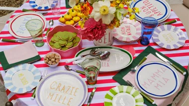 Table set with a red and white tablecloth, featuring In The Roundhouse Italian-style plates, along with a beautiful floral centrepiece. The spread includes knives, forks and cups,