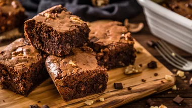 High angle view of various homemade chocolate brownie slices on a wooden cutting board surrounded by some brownie crumbs, some walnuts, a fork and a baking sheet with a baked brownie on the inside. Predominant colour is brown.