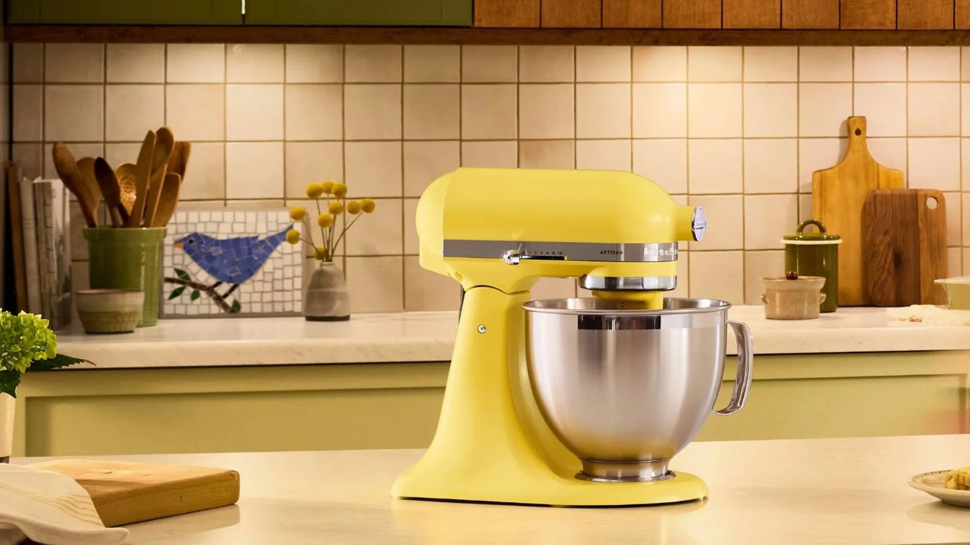 Yellow KitchenAid Artisan stand mixer with stainless steel bowl on a kitchen counter. Features a butter-coloured finish against a cream tile backsplash with wooden accessories in the background.