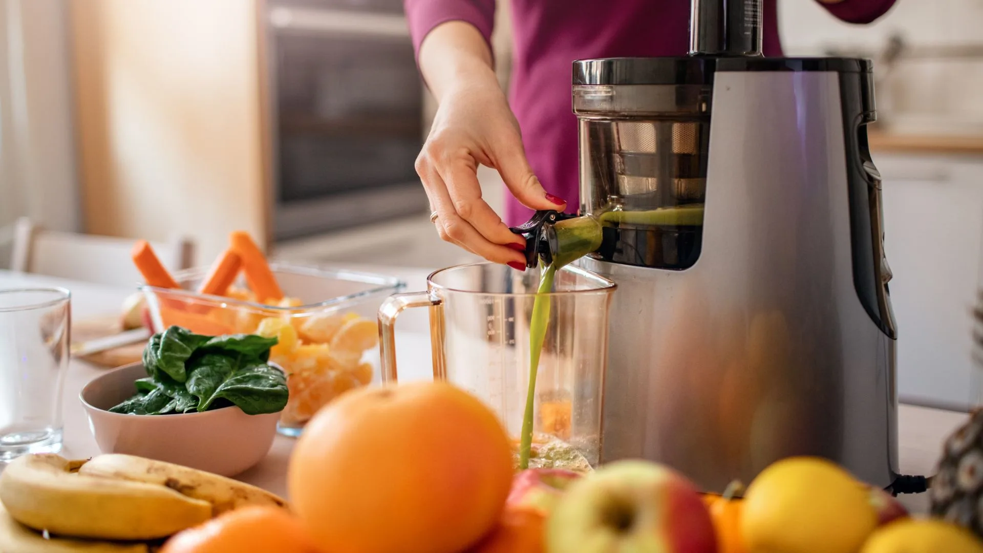 A person uses a cold-press juicer to make green juice with various fruits and vegetables on the counter.