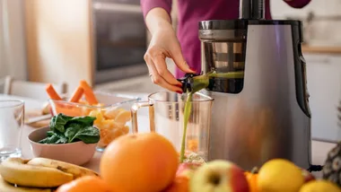 A person uses a cold-press juicer to make green juice with various fruits and vegetables on the counter.