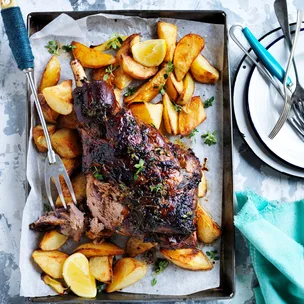 Greek roast lamb with skordalia and lemony potatoes on a baking tray. One slice of meat has been carved.