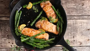Salmon fillets and asparagus spears cooking in a black Victoria seasoned cast iron skillet on a rustic wooden surface.