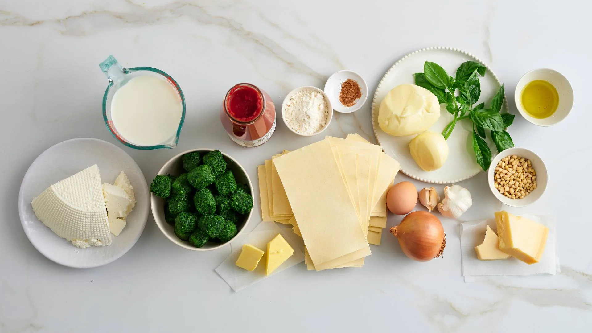 Spinach and ricotta ingredients flat lay of fresh ricotta, milk, frozen spinach, dried lasagne sheets, passata, garlic, nutmeg, parmesan, onions, basil, olive oil, pine nuts. 