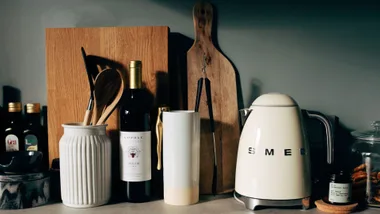 A stylish kitchen countertop displaying a collection of items including wooden chopping boards, a white ceramic utensil holder with wooden spoons, a bottle of red wine, a tall white and beige cylindrical container, black kitchen tongs, a cream-coloured Smeg electric kettle, and a small jar on a cork coaster.