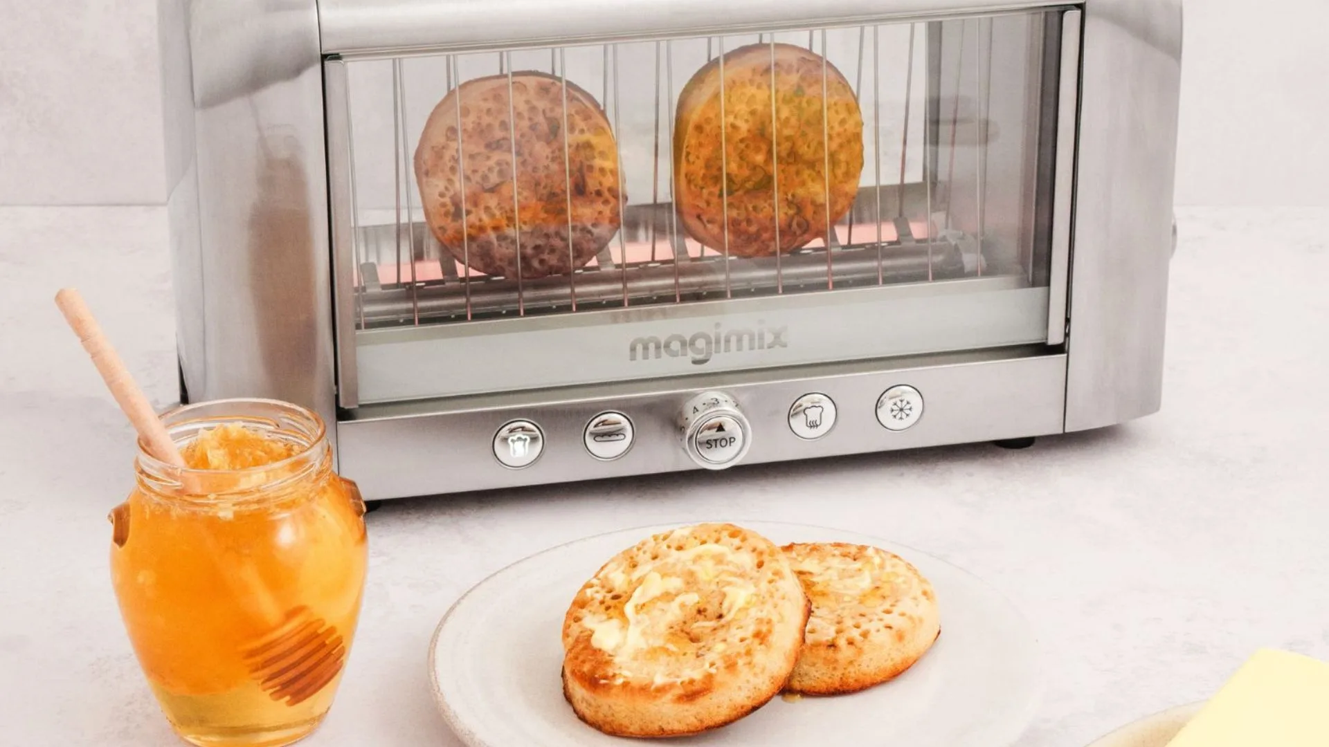 Magimix Vision Toaster with crumpets toasting inside, shown through the see-through glass panel. A jar of honey and a plate of buttered crumpets sit in front.
