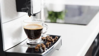 An image of a coffee machine, with a glass mug of coffee being dispensed and coffee beans scattered on the machine's tray.