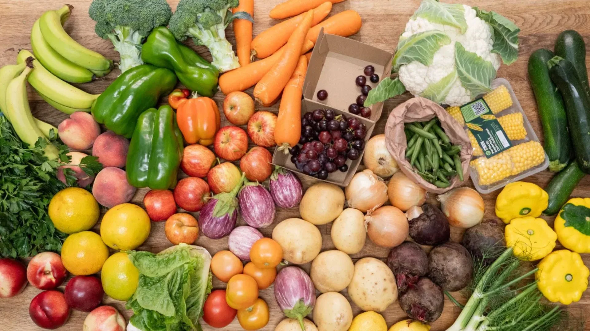 A colourful array of various green, orange, red, purple and yellow vegetables and fruits from Farmers Pick, spread out on a wooden table.