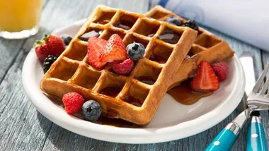 A stack of two square waffles drizzled with maple syrup and garnished with strawberries, blueberries, raspberries, and blackberries. The plate sits on a rustic blue wooden table next to a glass of orange juice.