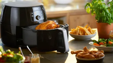 A Philips air fryer on a kitchen counter containing a whole roasted chicken. To the right are bowls of French fries (hot chips), roasted vegetables and sausages.