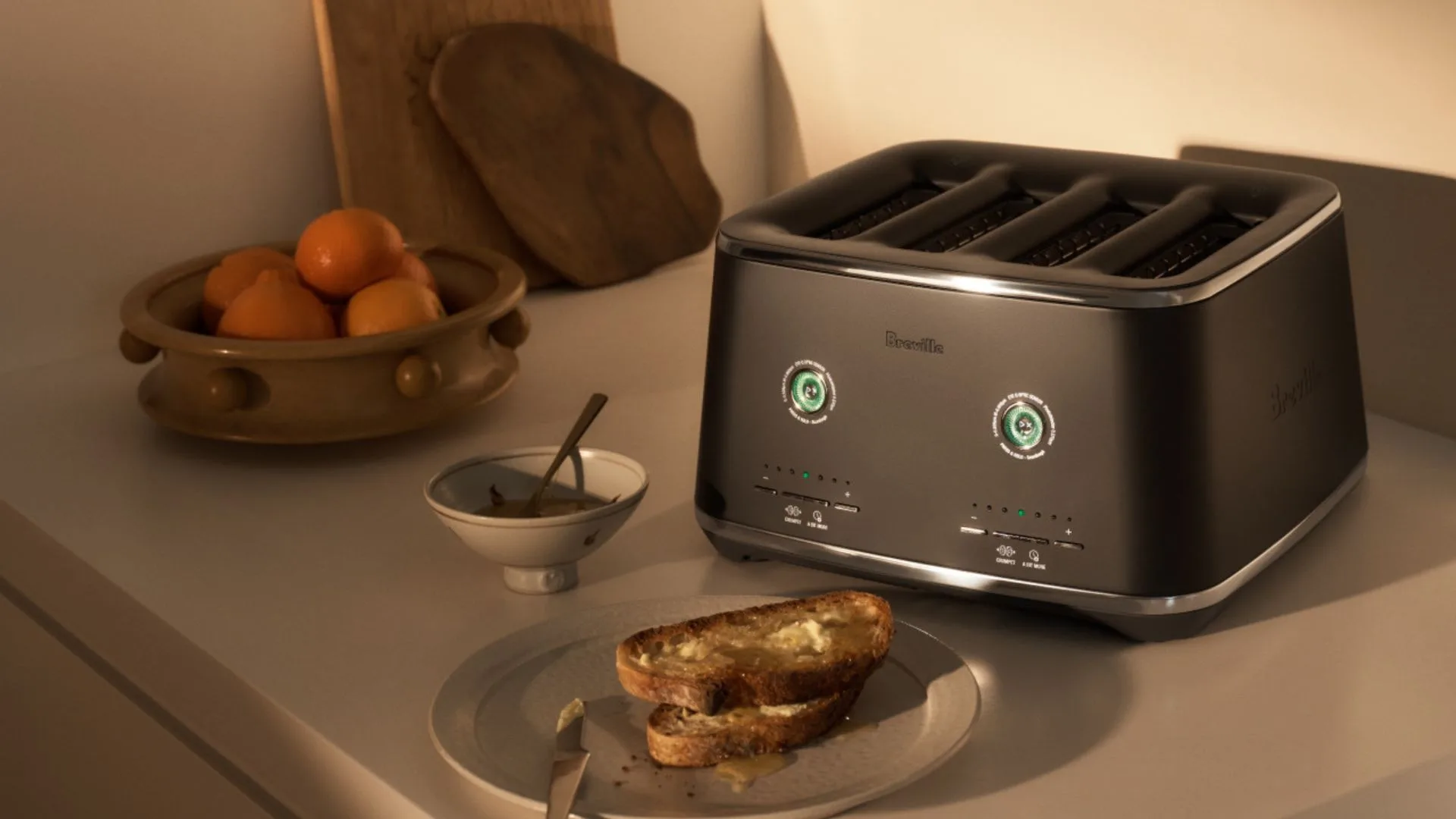A black Breville Eye Q Auto 4-slice toaster with silver accents sits on a kitchen countertop. The toaster's two main dials are lit up with green accents. In the foreground, there is a plate with two slices of buttered toast, and to the left, a bowl of oranges and a small dish with a spoon. The lighting is warm and golden.