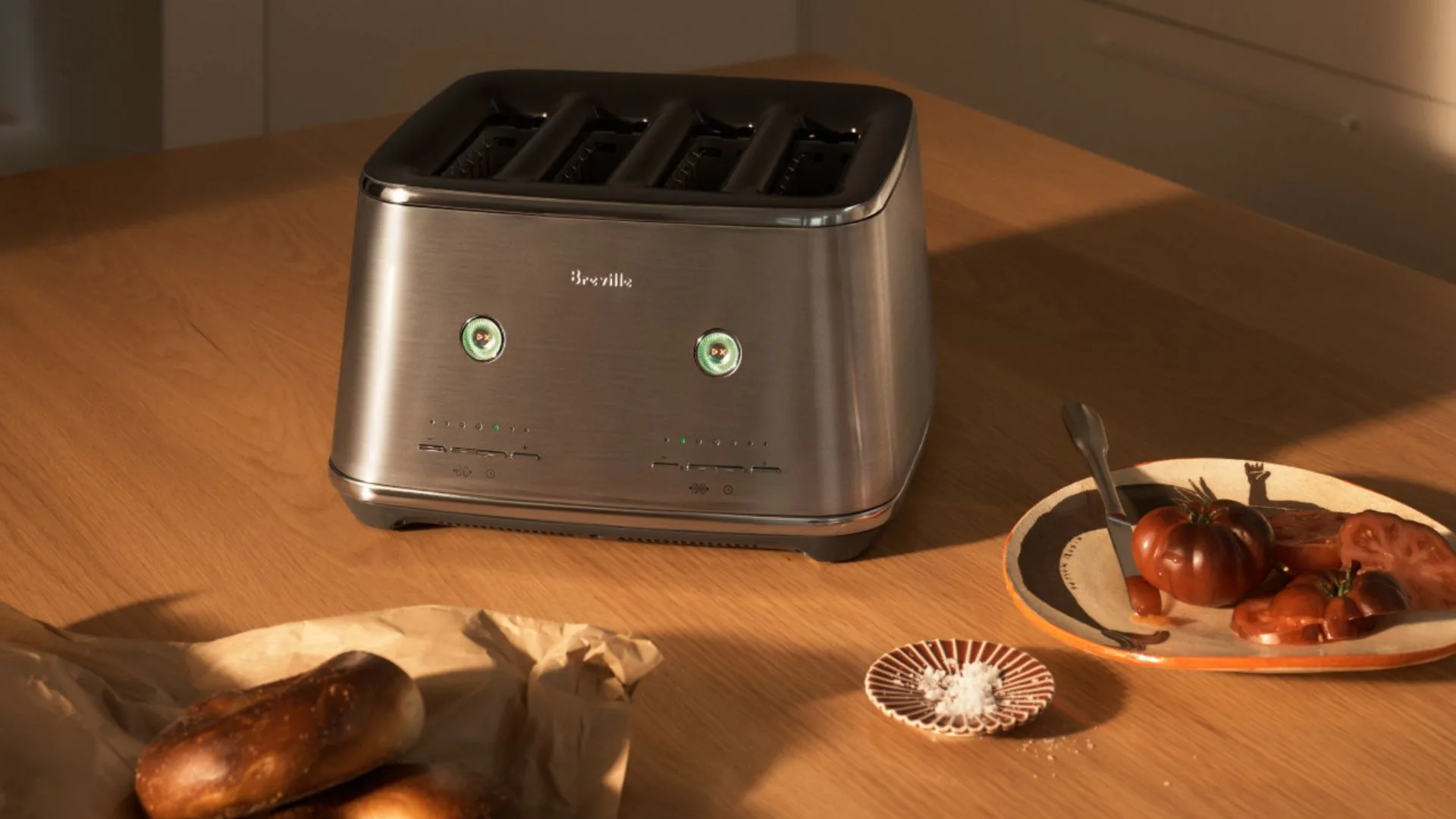 A Brushed Stainless Steel Breville Eye Q Auto 4-slice toaster sits on a wooden counter next to a plate of sliced heirloom tomatoes and some baked goods on a paper bag. The toaster's control dials are lit green.