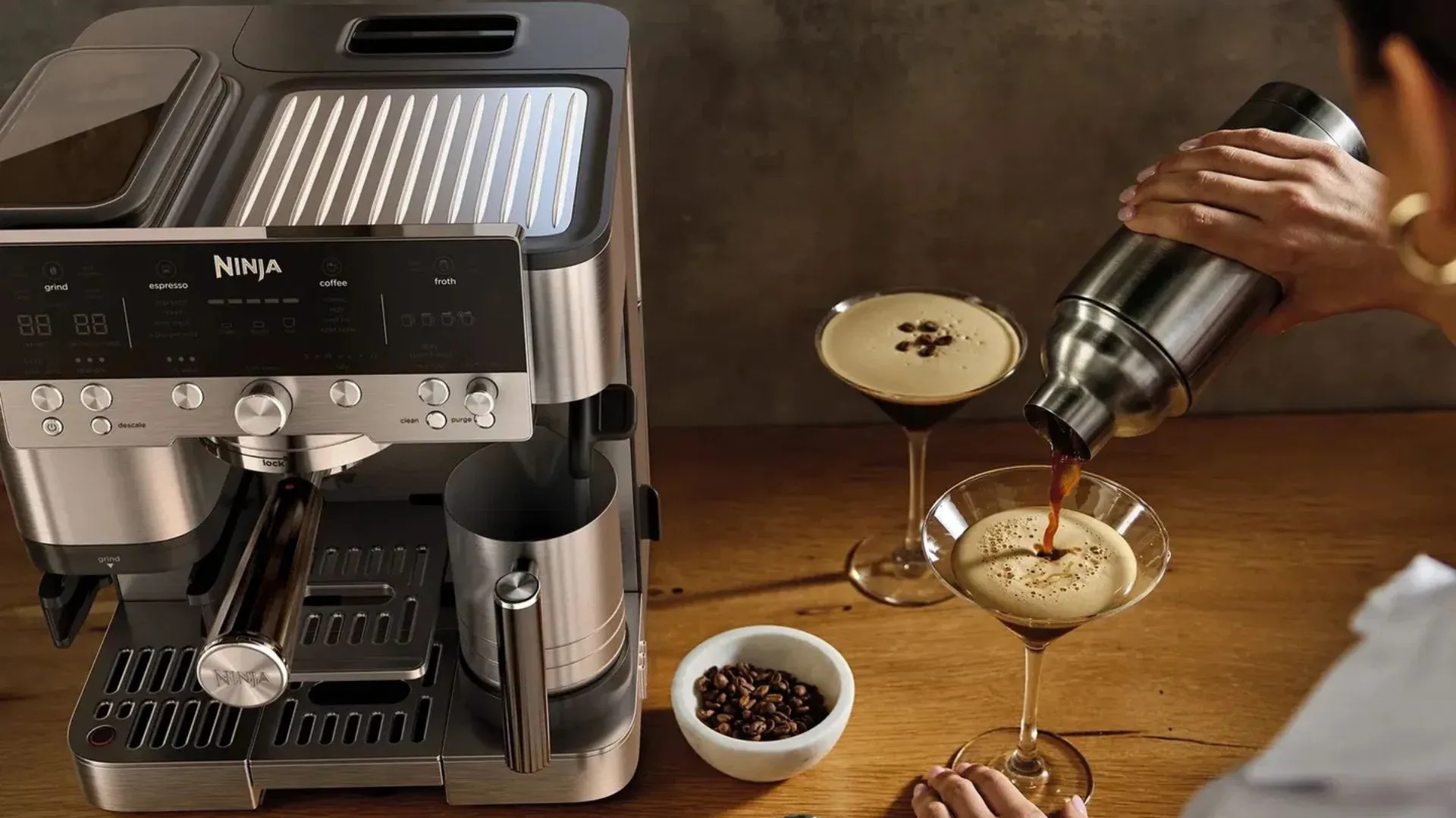 A close-up of the Ninja Café Deluxe coffee maker on the left. On the right, a person is shaking and pouring an espresso martini from a cocktail shaker into a martini glass. Another espresso martini and a small bowl of coffee beans are on the wooden countertop.