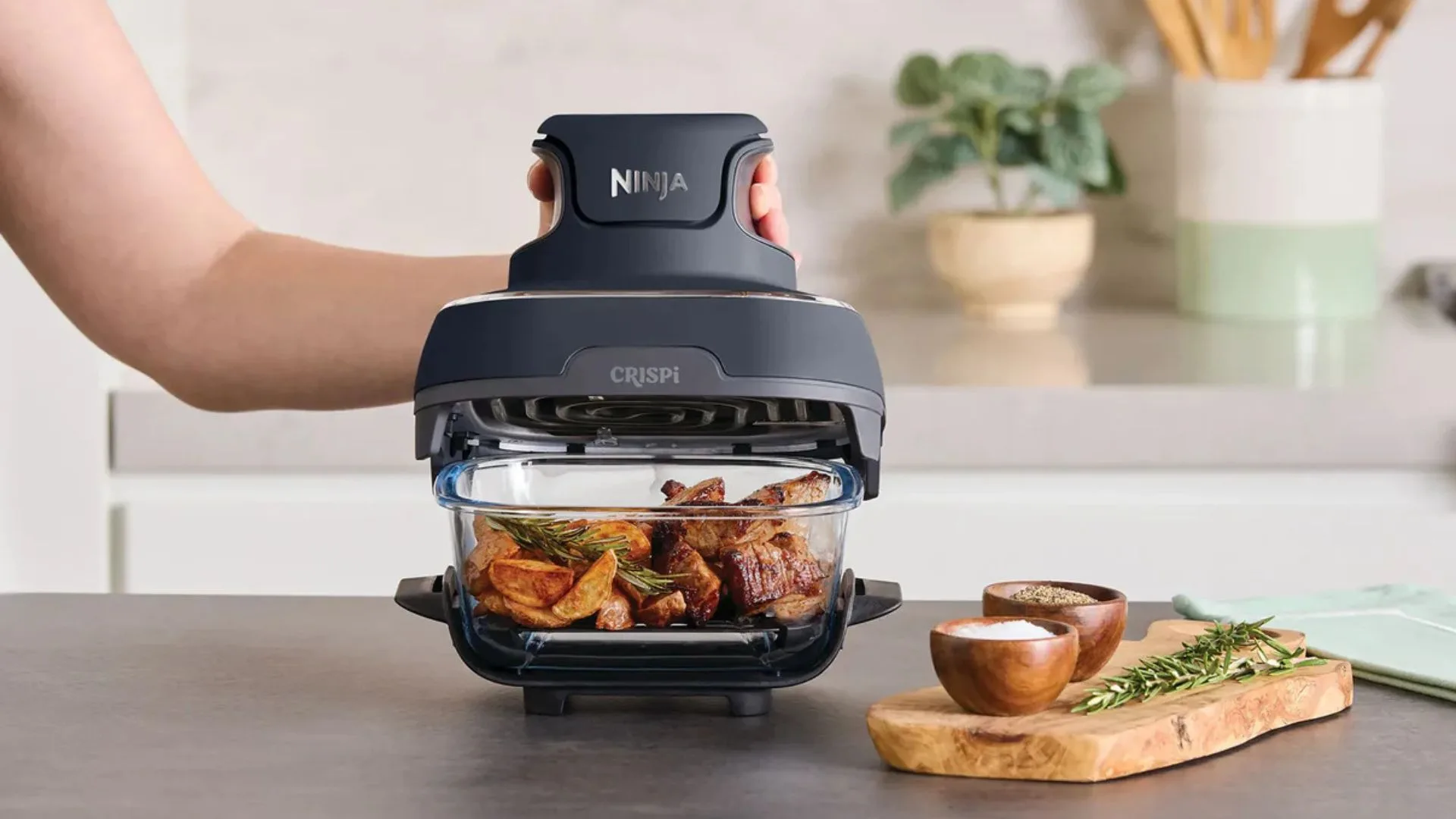 A close-up of the Ninja CRISPi air fryer in use, showing a hand lifting the dark grey main unit off the clear glass bowl. The bowl contains roasted meat and potatoes, sitting on a black base on a kitchen counter next to a wooden cutting board with salt, pepper and rosemary.