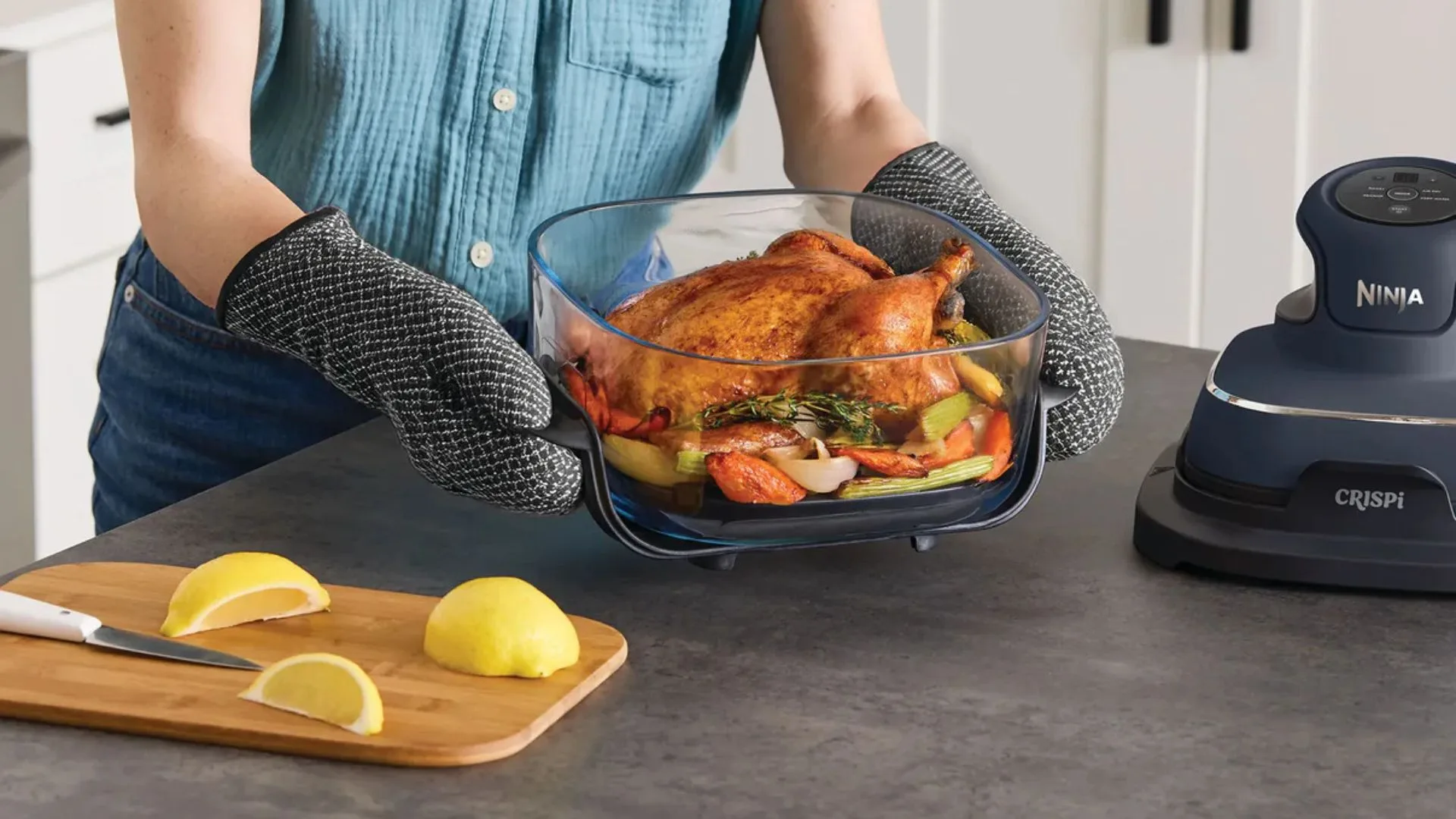 A person wearing black oven mitts lifts the clear glass bowl of the Ninja CRISPi air fryer, which contains a perfectly roasted whole chicken and vegetables. The dark grey air fryer base unit sits on the right, and a wooden cutting board with sliced lemons is on the left.
