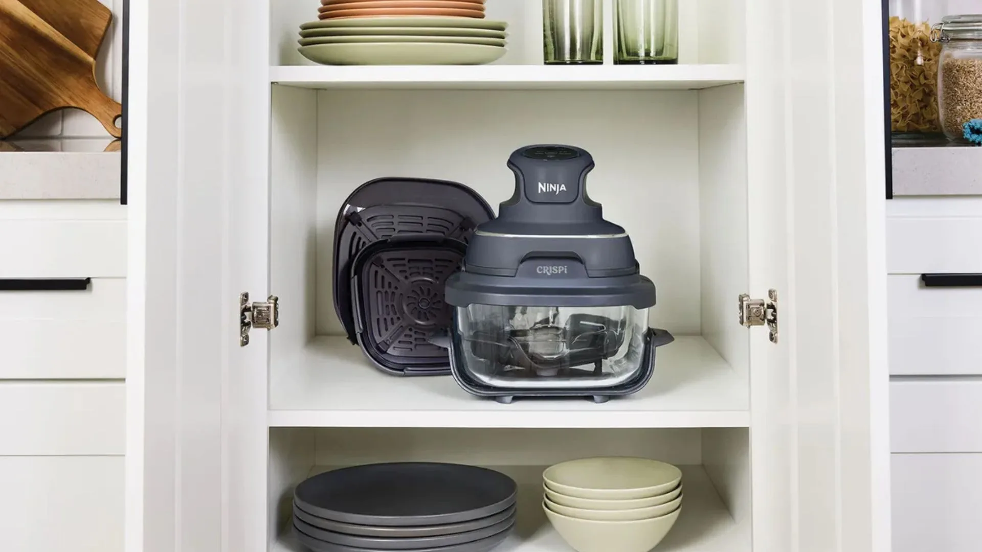 The compact Ninja CRISPi air fryer, dark grey with a clear glass bowl and two nested cooking trays, sits neatly on a white kitchen cupboard shelf surrounded by stacked dishes and bowls, illustrating its small footprint.