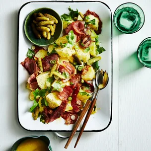Christmas ham and potato salad on a white platter with a bowl of cornichons and salad servers.