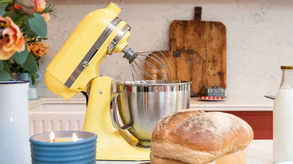 A KitchenAid stand mixer in the colour 'Butter' (yellow) sits on a kitchen counter next to fresh bread and pastries, with a whisk attachment in the stainless steel bowl.