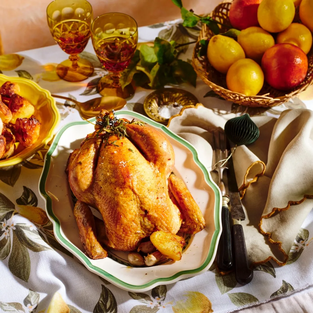 Maggie Beer roasted chicken and potatoes on serving dishes, with a bowl of fruit and two glasses.