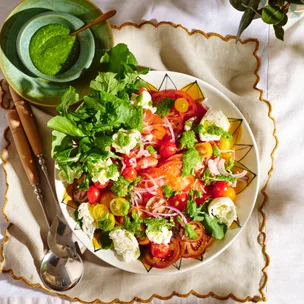 Maggie Beer's mozzarella and tomato salad recipe on a serving dish, with salad servers and a bowl of green pesto.