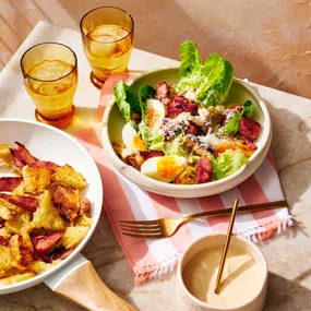 Leftover Christmas ham Caesar salad in a serving dish atop a striped tea towel, with a bowl of croutons and ham, plus a small bowl of dressing and two glasses of water.