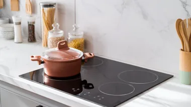 A copper cooking pot with a glass lid sitting on a modern black induction stovetop in a bright kitchen.