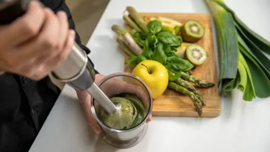 Close-up of a hand using an immersion blender to make a green smoothie, with fresh fruit and vegetables on a cutting board in the background.