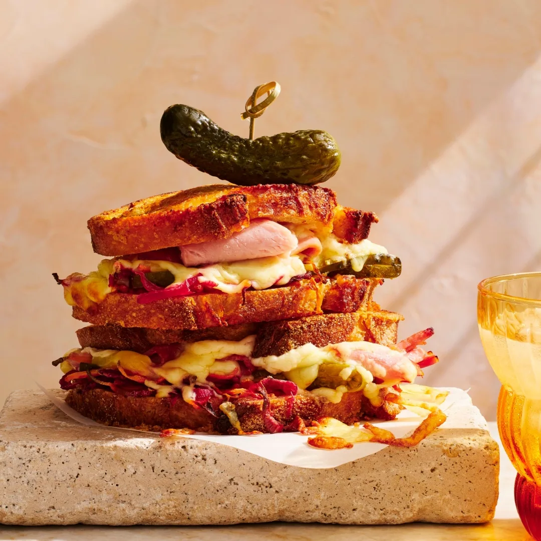 Toasted ham and cheese sandwich with a pickle on top, sitting on a stone slab.