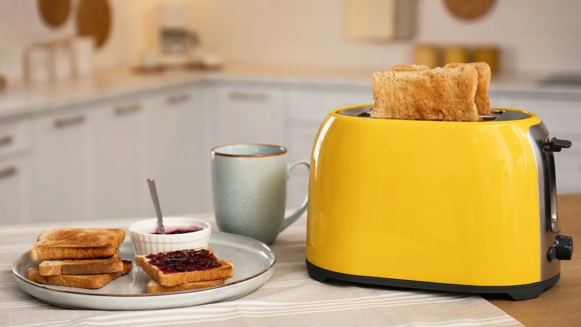 A bright yellow toaster with toast popping out, sitting on a table next to a plate of toast with jam and a blue mug.