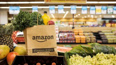 A brown paper grocery bag featuring Amazon and Harris Farm Markets logos, filled with fresh produce in a Harris Farm supermarket.