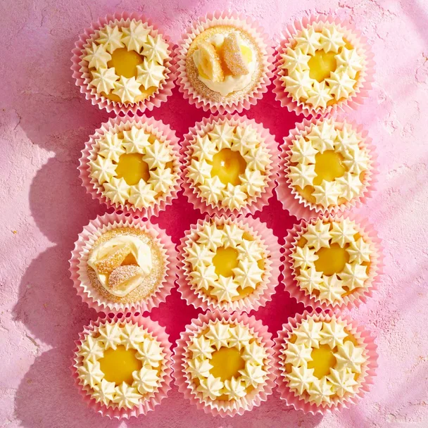 Twelve lemon curd cupcakes with pink patty cases on a pink surface.