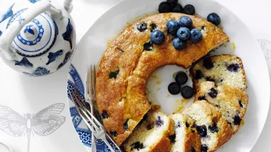A round vanilla cake with blueberries on a white platter and a pot of tea for morning tea