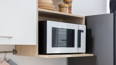 A sleek, modern white microwave with a black handle and trim, neatly tucked into a light-oak wood cabinet shelf.