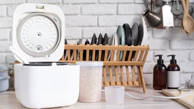 An open white rice cooker on a bright kitchen counter with a container of grains, a measuring cup and a wooden dish rack in the background.