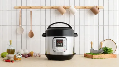 A stainless steel multi-cooker sitting on a kitchen benchtop surrounded by fresh cooking ingredients like garlic, onions, and herbs.
