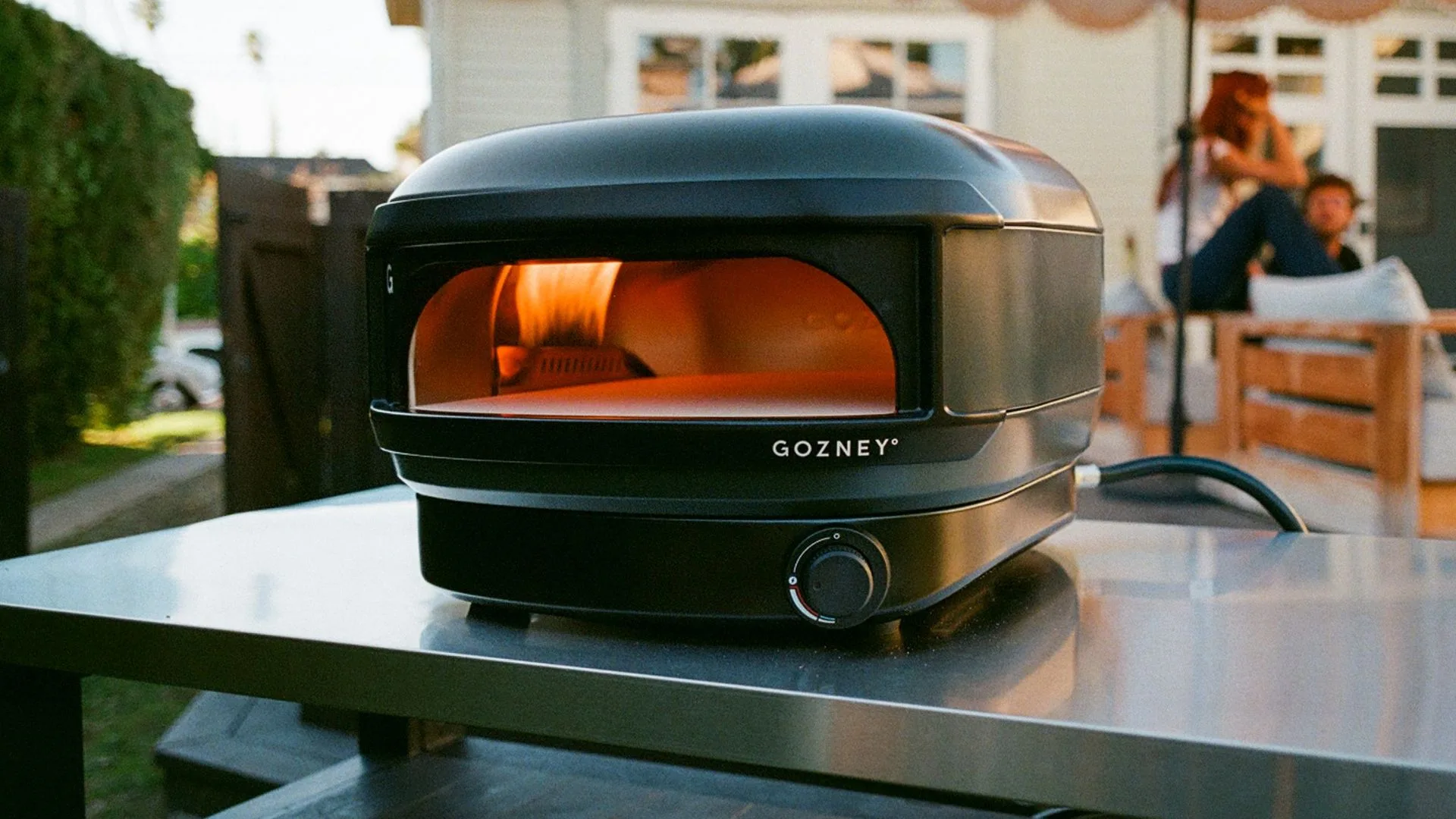 A close-up shot of the Gozney Arc Lite pizza oven in Off Black sitting on a stainless steel outdoor bench, showing the glowing lateral flame and the compact, modern design.